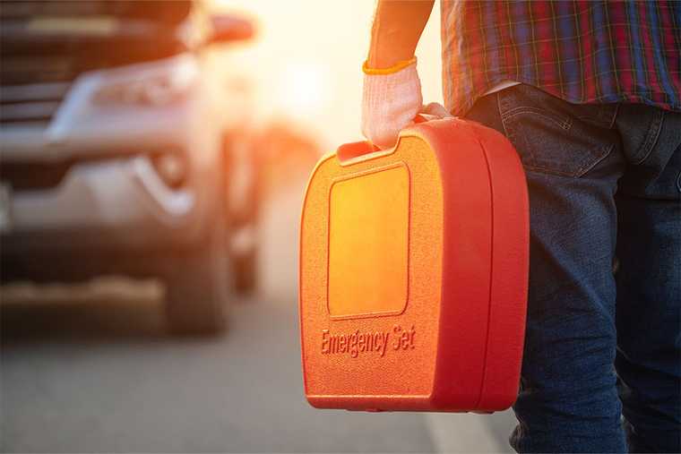 A person carrying an orange plastic case labeled "Emergency Set" toward a blurred vehicle parked on the side of a road.