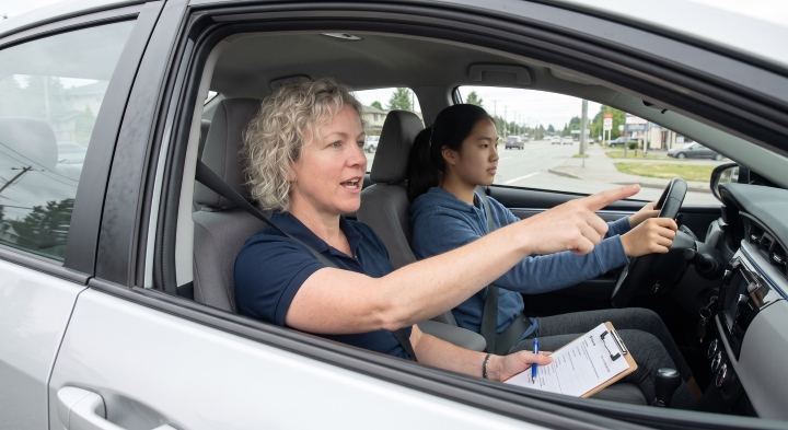 driving instructor sitting in the passenger seat next to a driver inside a car, with the Instructor pointing ahead and talking.