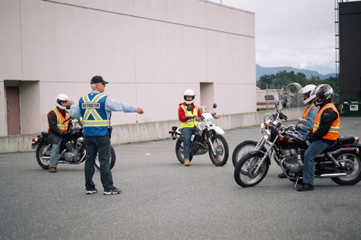 A motorcycle instructor wearing a high-visibility vest standing amongst four students on motorcycles, directing the students.