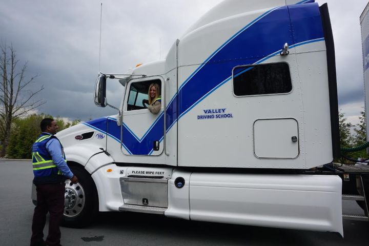 A side profile of a white semi-truck with blue accent stripes parked outside, with a student in the driver's seat and the Instructor outside the truck.