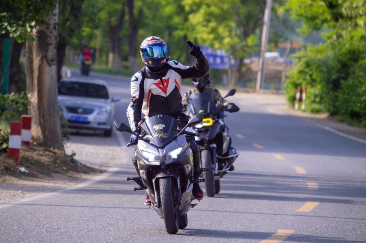 Two motorcycle riders in full gear riding their motorcycles side-by-side along a paved road, with one of them gesturing a peace sign.