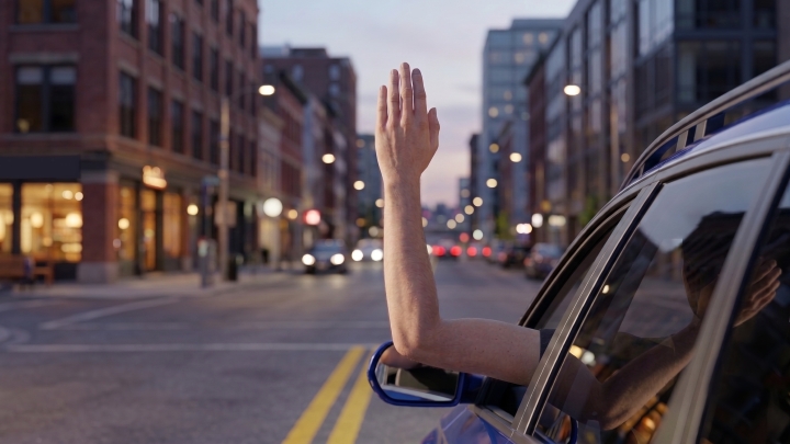 A view of a driver extending their left arm out of a car window, bent upward at a 90-degree angle at the elbow to make a right-turn hand signal.