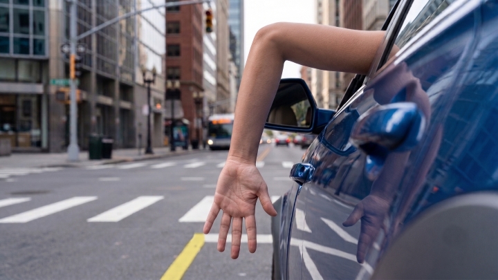 A view of a driver extending their left arm out of a car window, bent downward at a 90-degree angle at the elbow with the palm facing backward to signal stopping or slowing down.