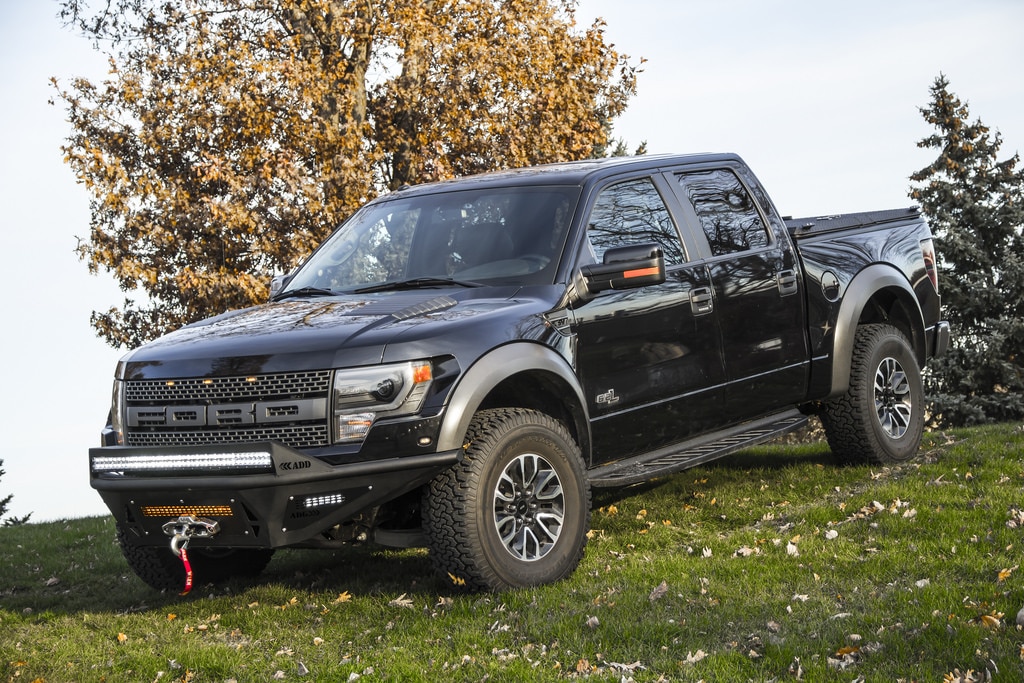 A black Ford F-150 Raptor with an off-road front bumper and LED light bar, parked on a grassy field with autumn leaves and trees in the background.