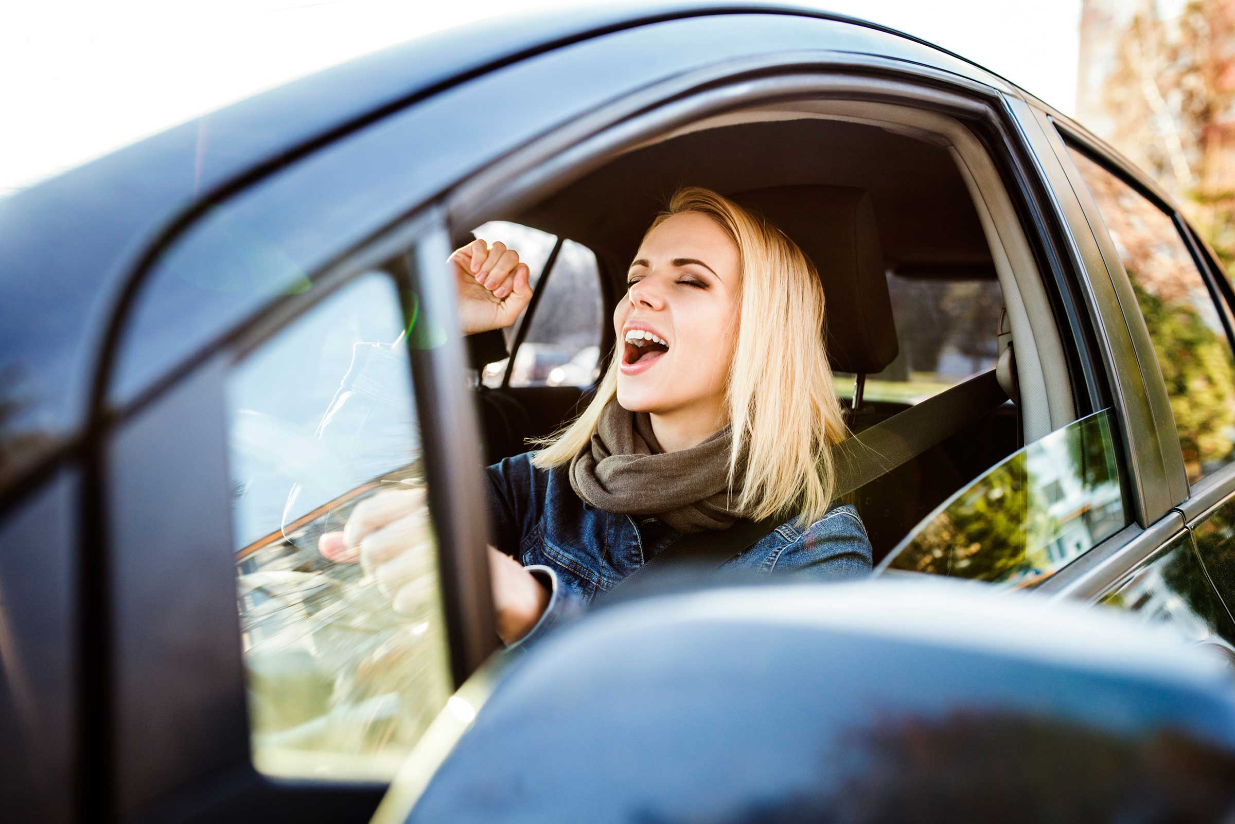 A woman with long blonde hair leaning out of a car window with her eyes closed and mouth open, looking as though she is singing or cheering.