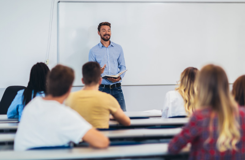 An instructor standing at the front of a classroom holding a book and speaking to a group of seated students.