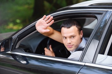 A man sitting in the driver's seat of a car, looking back and waving his hand out of the open window.