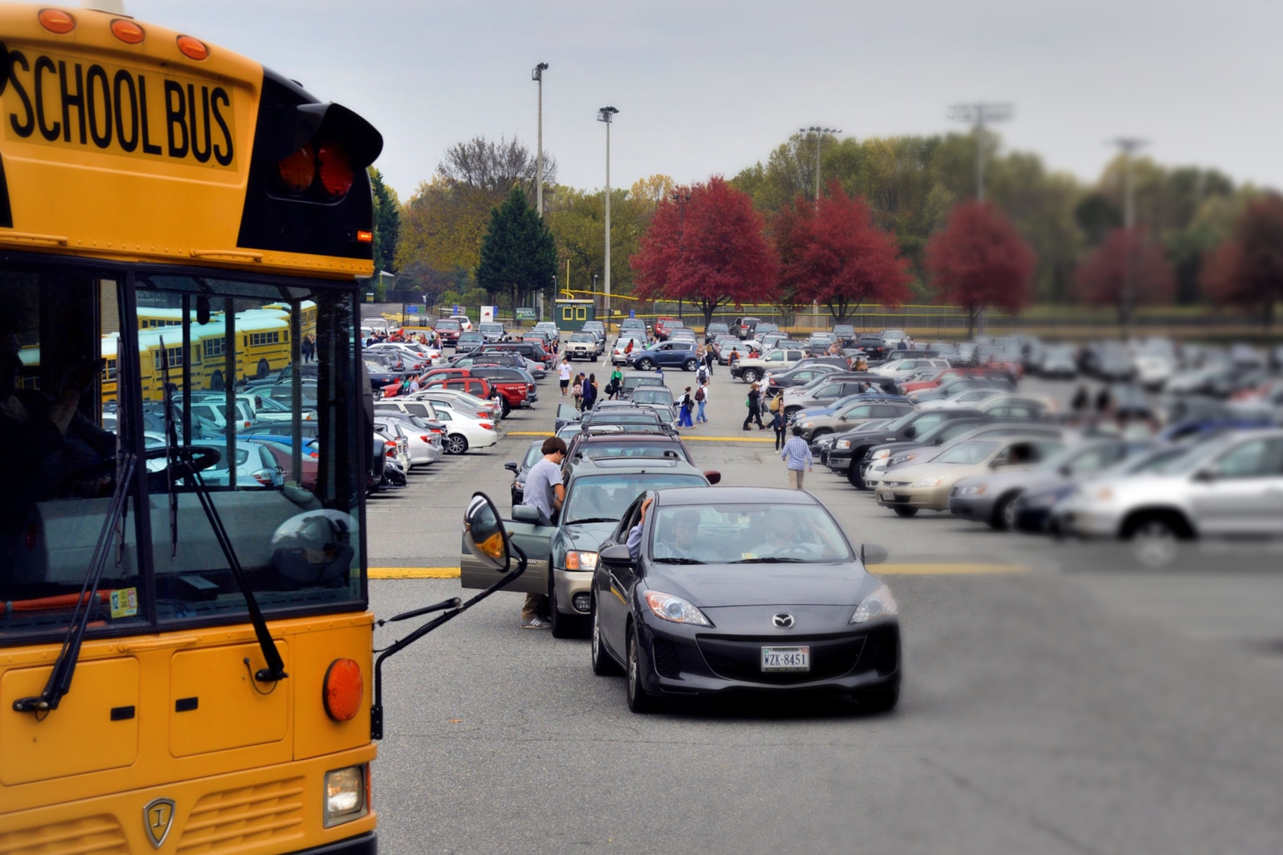 A yellow school bus in the foreground next to a crowded parking lot filled with cars and people.