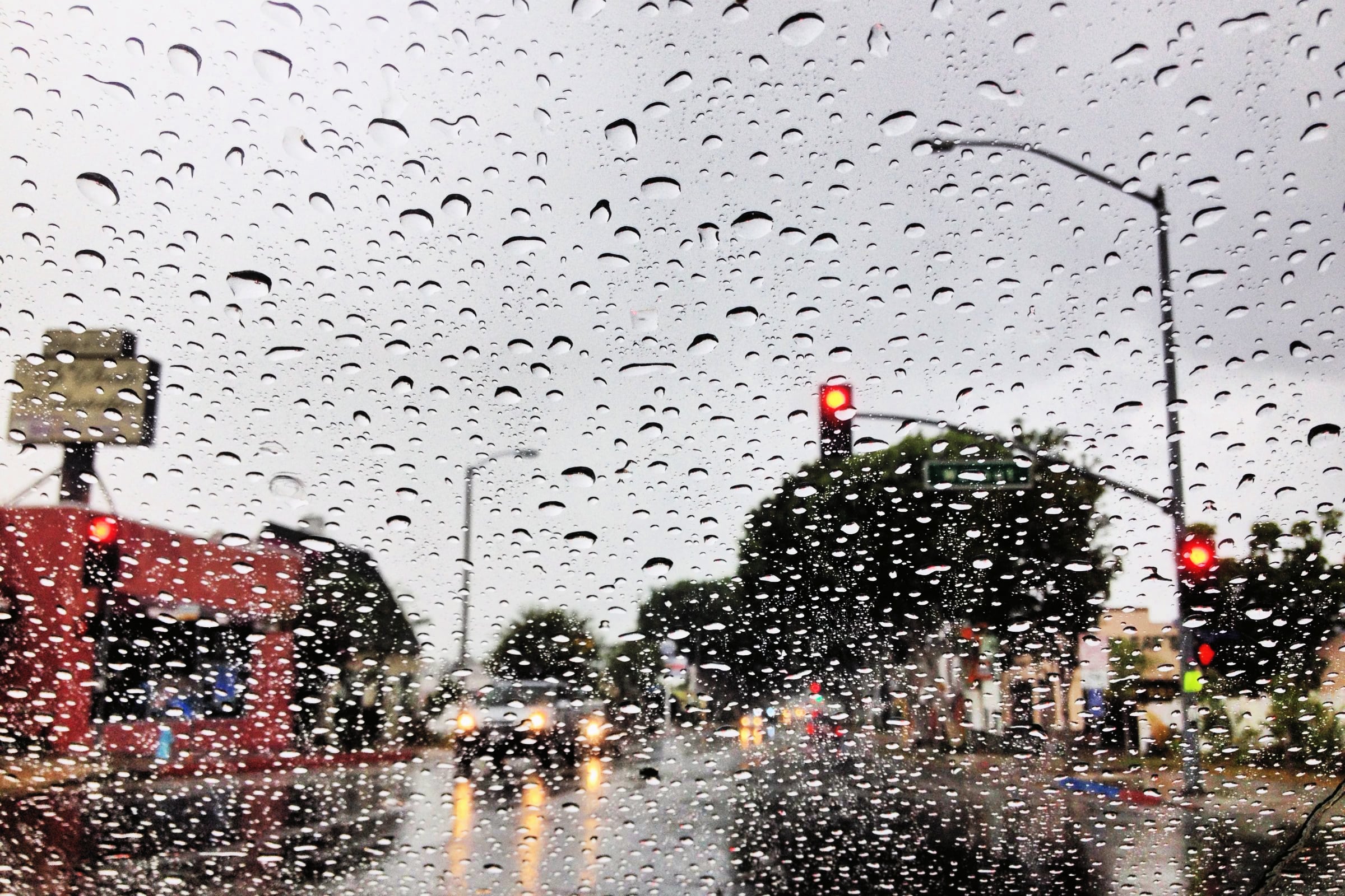 A view through a rain-streaked windshield of a city street with red traffic lights and blurred car headlights at dusk.
