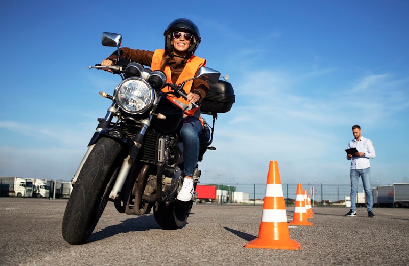 A female motorcyclist wearing a helmet, sunglasses, and an orange safety vest, smiling while sitting on a motorcycle; an instructor with a clipboard stands in the background near orange traffic cones.