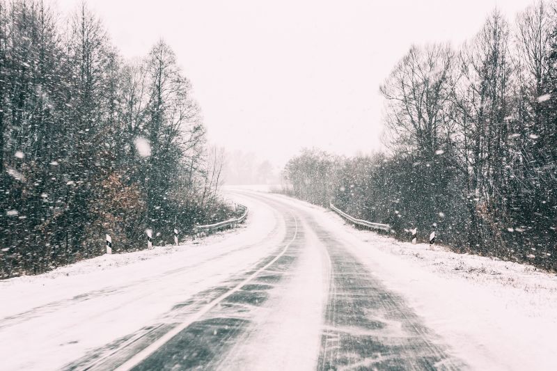 A winding two-lane road covered in snow and ice passes through a wintry forest of bare trees, with large snowflakes falling across the scene.