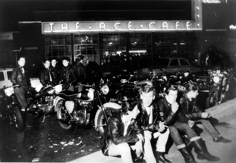 A black-and-white historical photo of a group of people in leather jackets gathered with their motorcycles in front of "THE ACE CAFE" at night.
