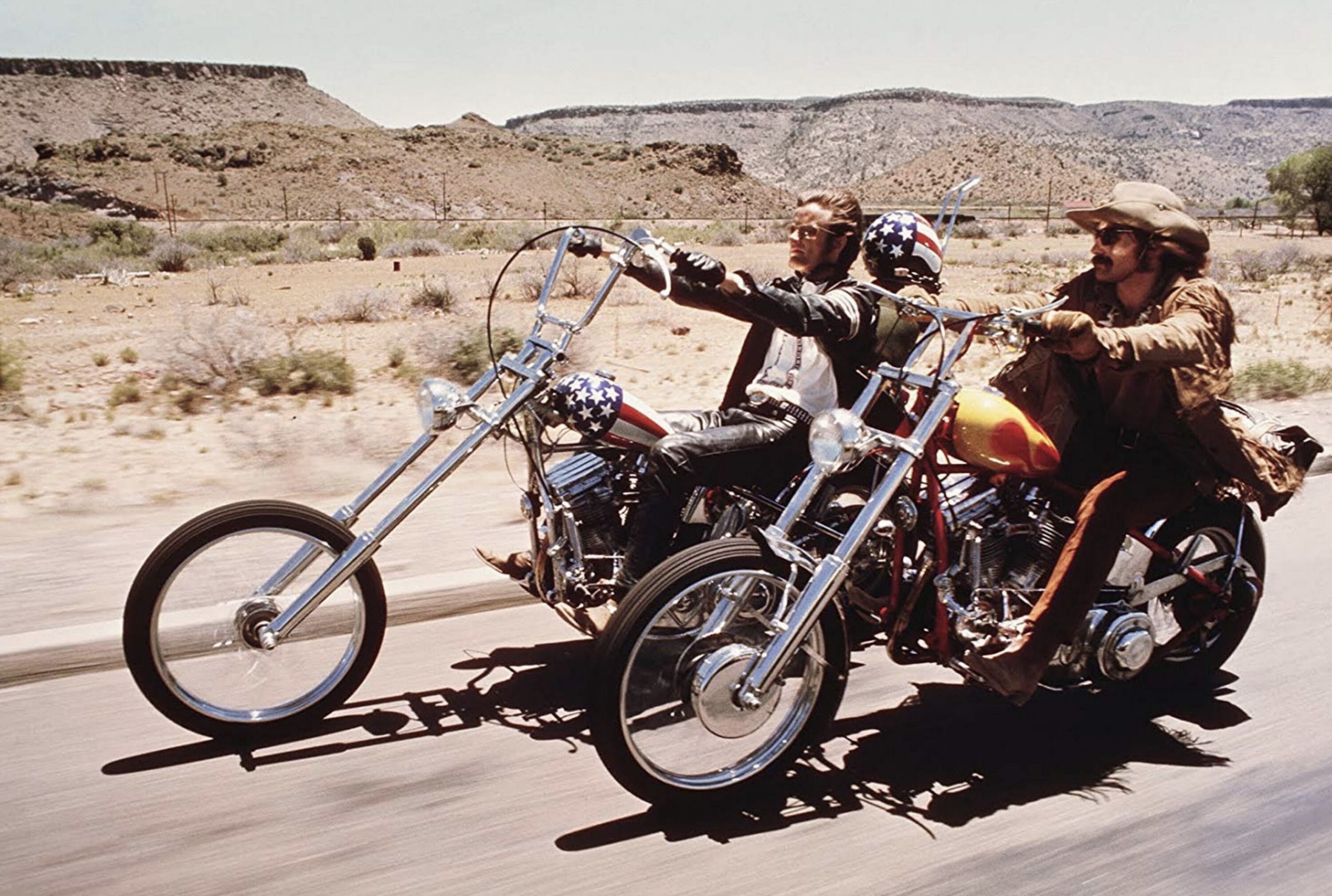Two men riding custom chopper motorcycles side-by-side on a desert highway.