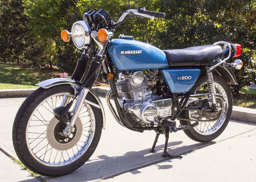 A blue vintage Kawasaki KZ200 motorcycle parked on a sidewalk with green trees in the background.