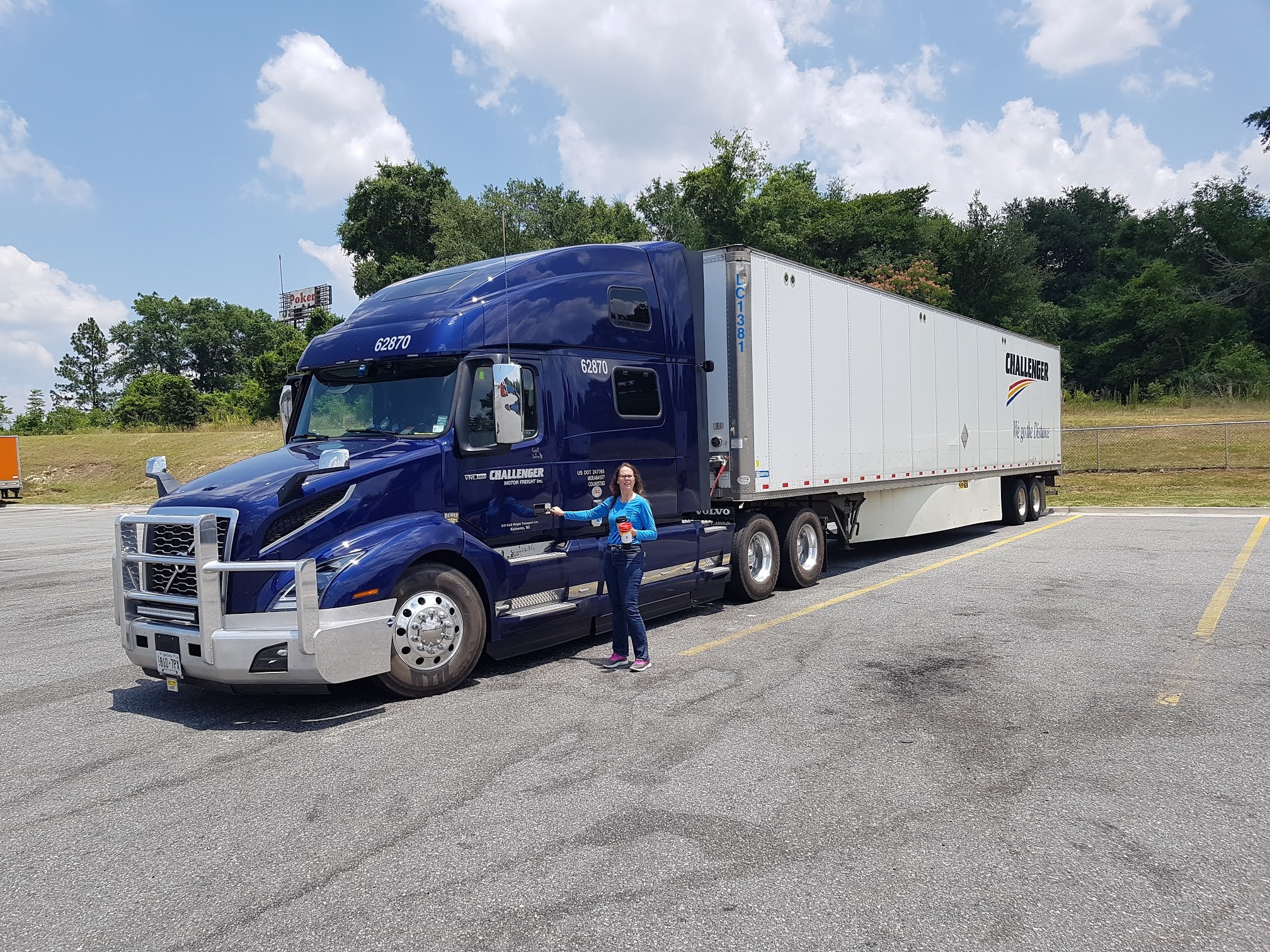 Woman in a blue shirt standing beside a large blue Volvo tractor-trailer in a parking lot on a sunny day with a white trailer attached