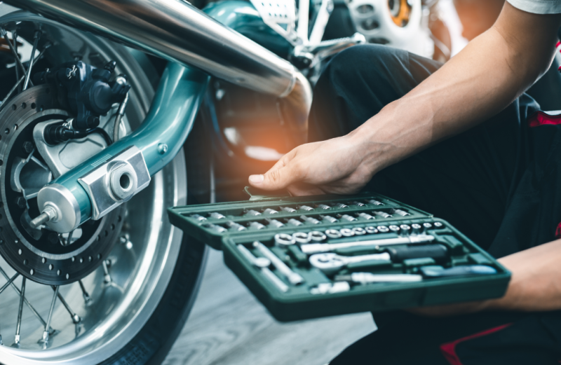 A close-up view of a mechanic's hand holding a tool kit, with a motorcycle in the background.