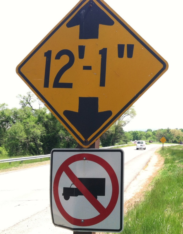 A yellow diamond-shaped sign indicating a height clearance of 12 feet 1 inch, mounted above a white square sign with a red "no trucks" symbol on a roadside.