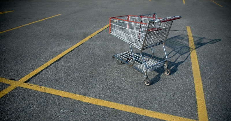 A single, empty metal shopping cart left in the middle of an asphalt parking lot within yellow-lined parking stalls.