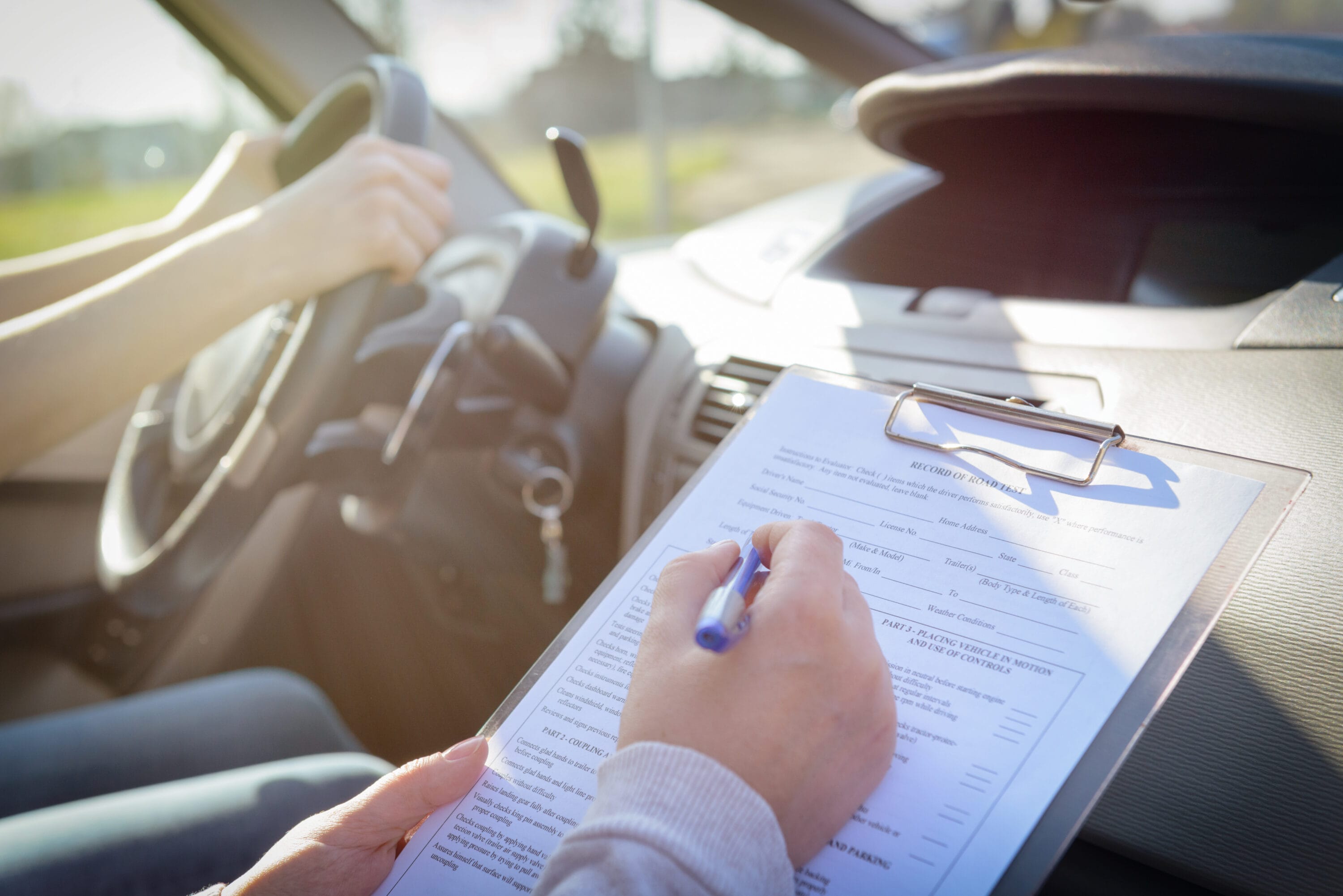 zoomed in on 2 people in a car with a driver holding the steering wheel and the passenger writing on a clipboard.