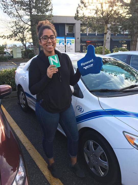 A student smiling and holding an I Passed sign while standing next to a Valley Driving School car.