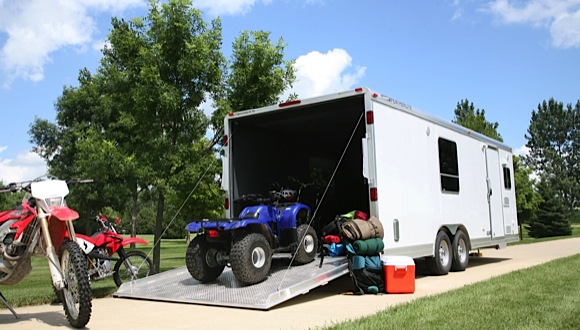 A white enclosed toy hauler trailer with its rear ramp lowered, showing a blue ATV being loaded and a red dirt bike and camping gear stacked nearby.