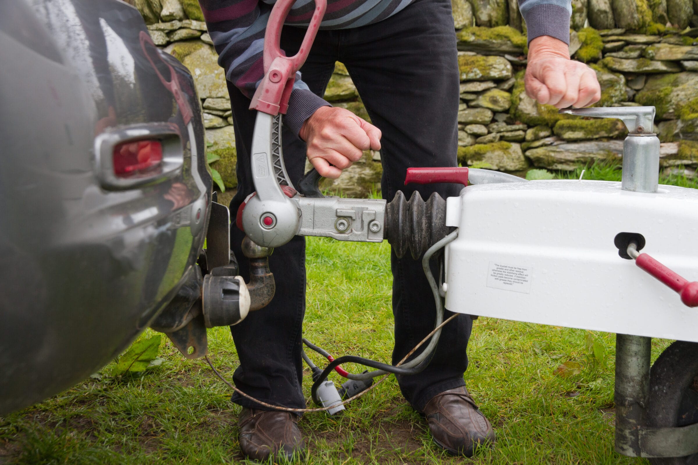 A person's hands adjusting the hitching mechanism of a white trailer to connect it to the tow ball of a grey car.