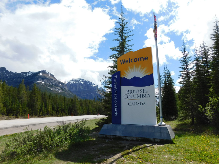 A large "Welcome to British Columbia, Canada" sign standing alongside a highway, featuring the slogan "The Best Place on Earth" and a backdrop of dense evergreen forests and majestic, snow-capped mountains.