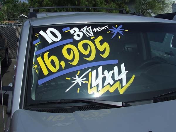 A silver SUV on a car lot with "16,995", "3RD SEAT", and "4X4" painted in bright yellow and white on the windshield.