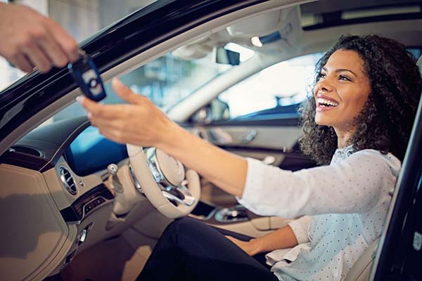 A smiling woman sitting in the driver's seat of a new car while reaching out to receive a set of keys from someone outside the vehicle.