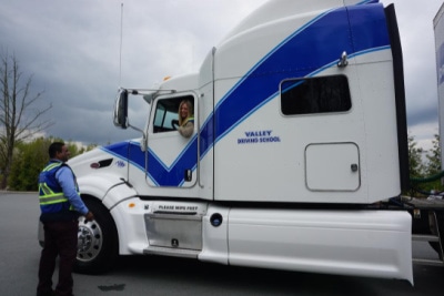 An instructor in a neon safety vest stands outside a white Valley Driving School semi-truck, gesturing and talking to a student driver sitting in the cab.