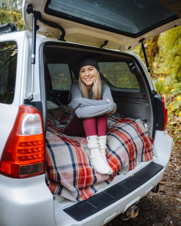 A smiling young woman sits comfortably on a red and grey plaid blanket inside the open trunk of a white SUV, dressed in a beanie and cozy winter clothing while parked in a forest.
