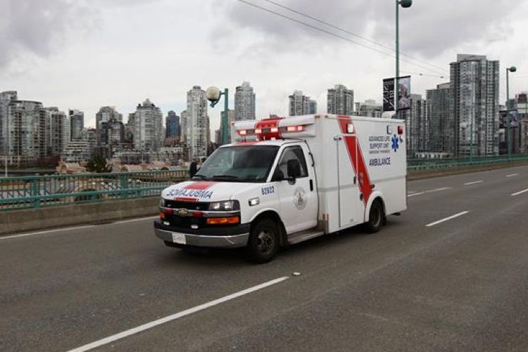 A white Advanced Life Support ambulance with red accents and flashing emergency lights drives across a multi-lane bridge, set against a backdrop of a dense urban skyline under an overcast sky.