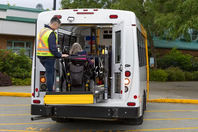 A HandyDART driver assisting a passenger using the rear hydraulic lift of a shuttle bus.