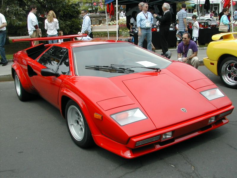A red Lamborghini Countach parked on a paved surface with several people and other vehicles in the background.