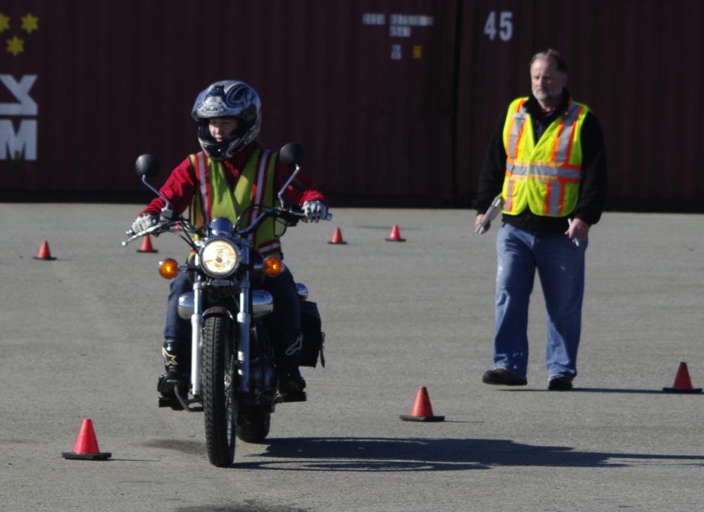A person riding a motorcycle through an obstacle course of orange cones while an instructor in a safety vest watches.