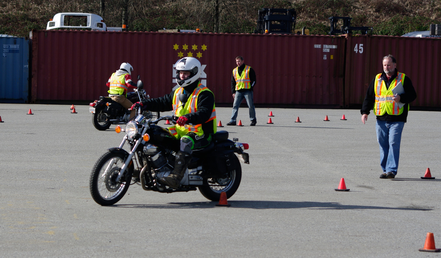 Two motorcycles riding in a parking lot and navigating orange traffic cones with 2 Instructors in safety vests observing.