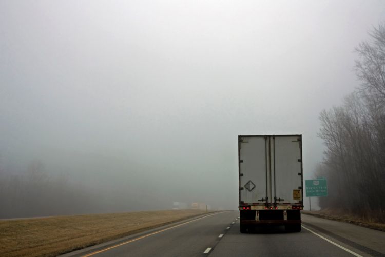 The rear of a white semi-truck traveling on a highway as it enters a thick, grey wall of fog, with bare trees and a green highway sign partially obscured on the roadside.