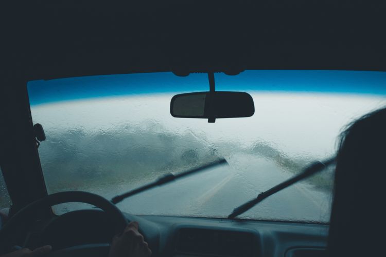 A view from the interior of a car looking out through a rain-streaked windshield during a heavy downpour, with the windshield wipers in mid-motion and the road ahead barely visible.