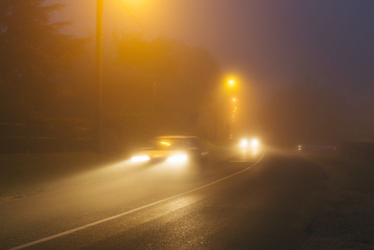 A nighttime scene on a dark road where thick, heavy fog severely limits visibility, with the bright beams of oncoming car headlights and the warm orange glow of streetlights cutting through the dense mist.