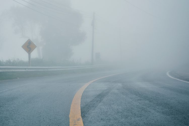 A paved road with a yellow center line curves into a wall of extremely dense white fog, with a yellow winding road sign and metal guardrail barely visible on the left.