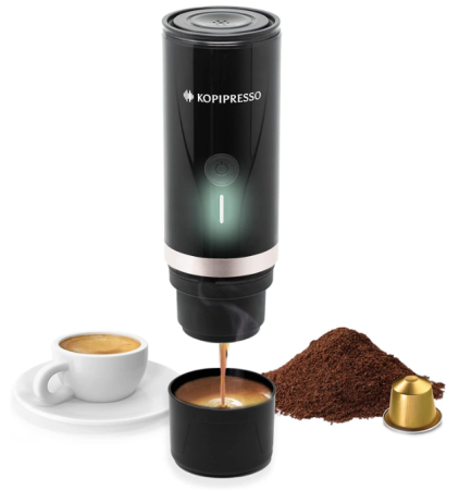 A black portable espresso maker standing next to a small glass of espresso and a pile of coffee beans.