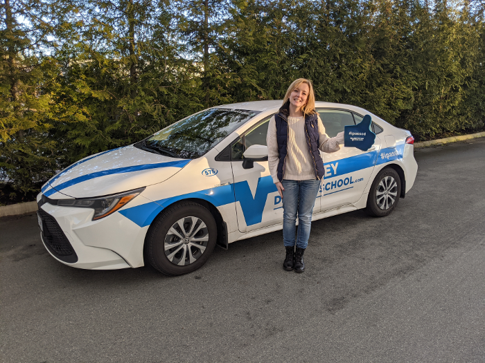 a young woman stands in front of a Valley Driving School car holding a sign that says "I passed".