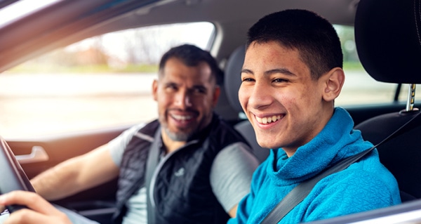 2 men in a car. The driver is a young man who is smiling and there is an older man, also smiling, in the passenger seat.
