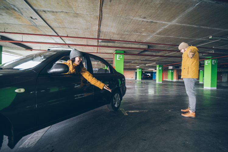 A person in a yellow jacket standing in a parking garage, watching a driver in a black car attempt to park in a tight space.