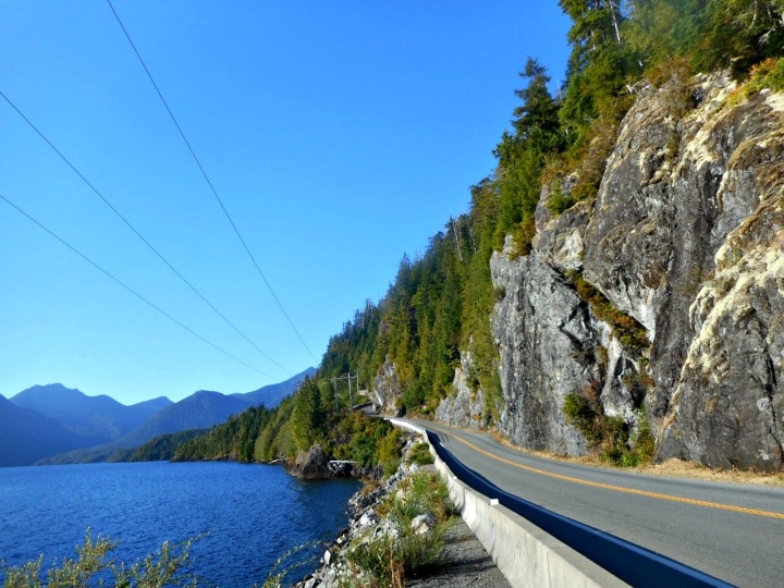 A paved highway curving along a rocky cliffside next to a large blue lake, with mountains in the background under a clear sky. Vancouver Island, BC.