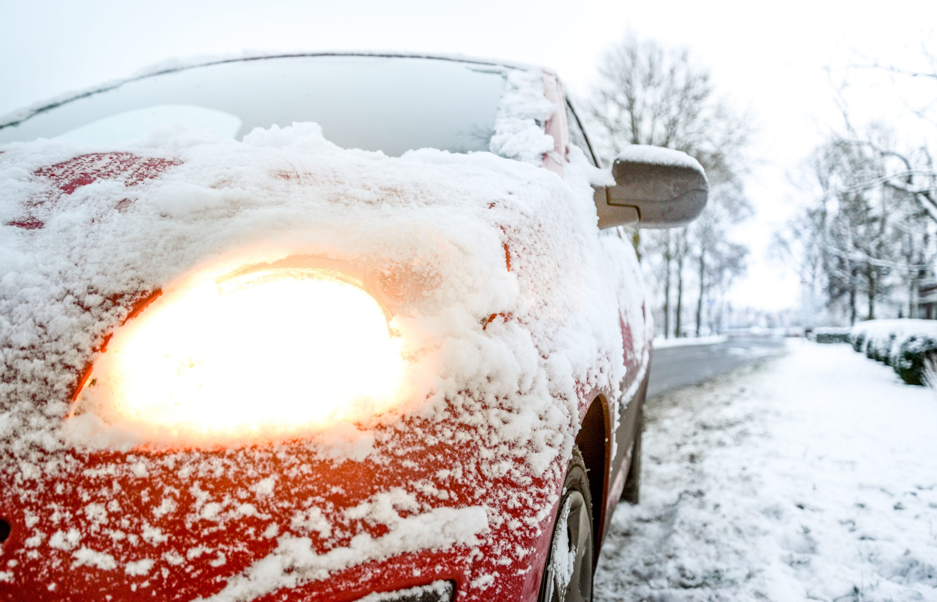 A close-up of a red car covered in snow with its headlight turned on, parked along a snow-lined street.