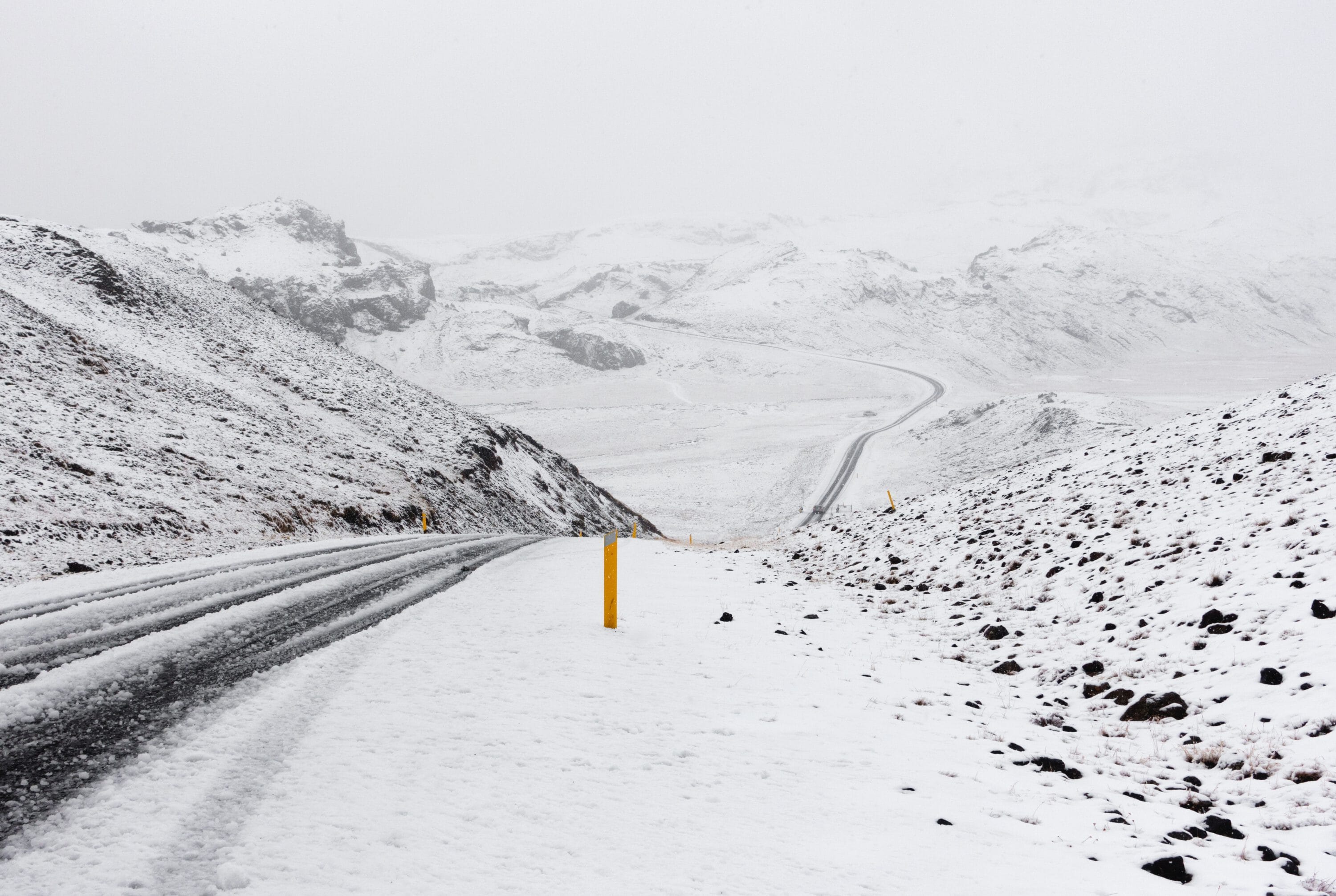 A winding, slushy road cutting through a vast, snow-covered mountain landscape under a cloudy sky, with yellow markers along the shoulder.
