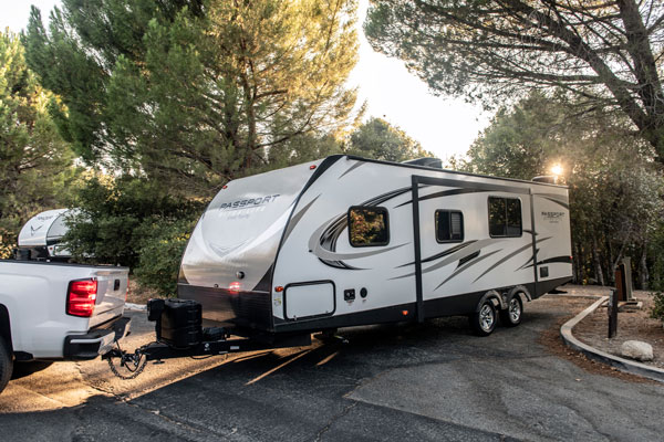 A white pickup truck hitched to a large white and grey Passport travel trailer, parked on a paved surface in a wooded area with sunlight filtering through the trees.