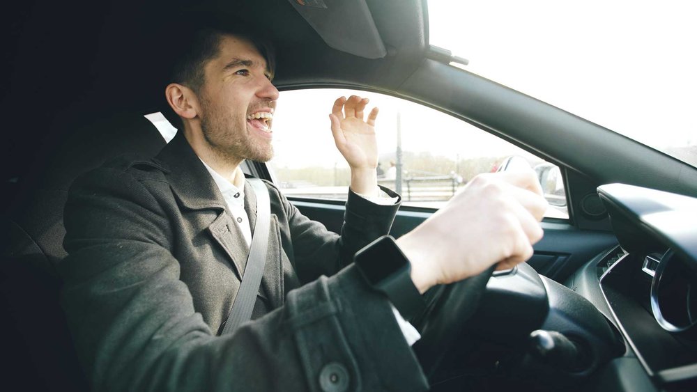 A man in a grey jacket singing enthusiastically while sitting in the driver's seat of a car.