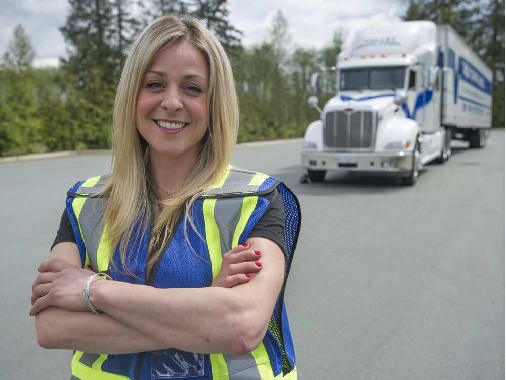 A woman in a blue safety vest stands in a parking lot in front of a semi truck with her arms folded.